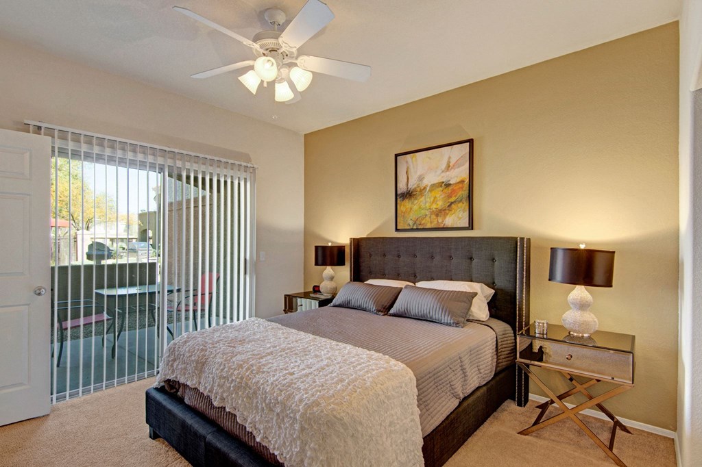 Modern bedroom here at Dobson Towne Centre Apartments with a gray upholstered bed, striped bedding, and a textured white throw. Warm lighting from side lamps, abstract art above bed, and sliding glass door with vertical blinds leading to a patio.