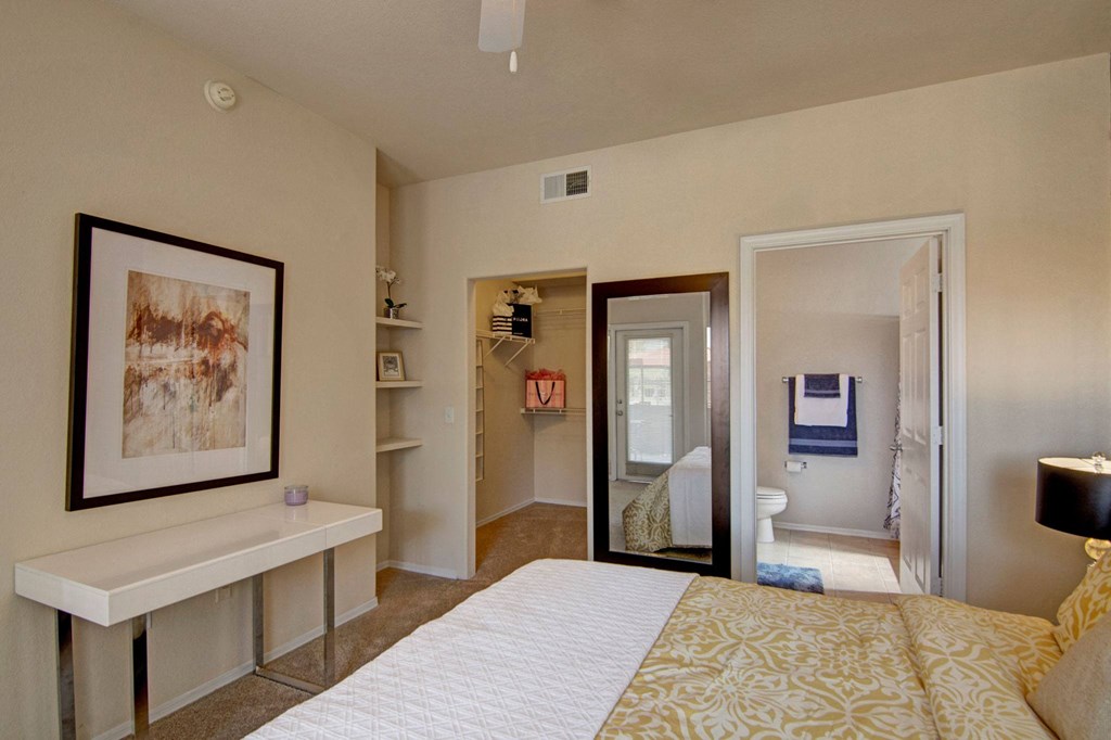Modern bedroom here at Dobson Towne Centre Apartments with beige walls, white desk, abstract art, and a large mirror. Walk-in closet and bathroom visible, creating a cozy, organized feel.