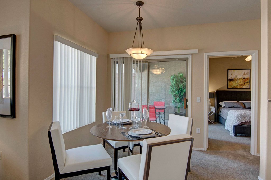 Dining area here at Dobson Towne Centre Apartments with a round table set for four. White chairs and a modern chandelier create a cozy atmosphere. A glass door leads to a patio, and a bedroom is visible.