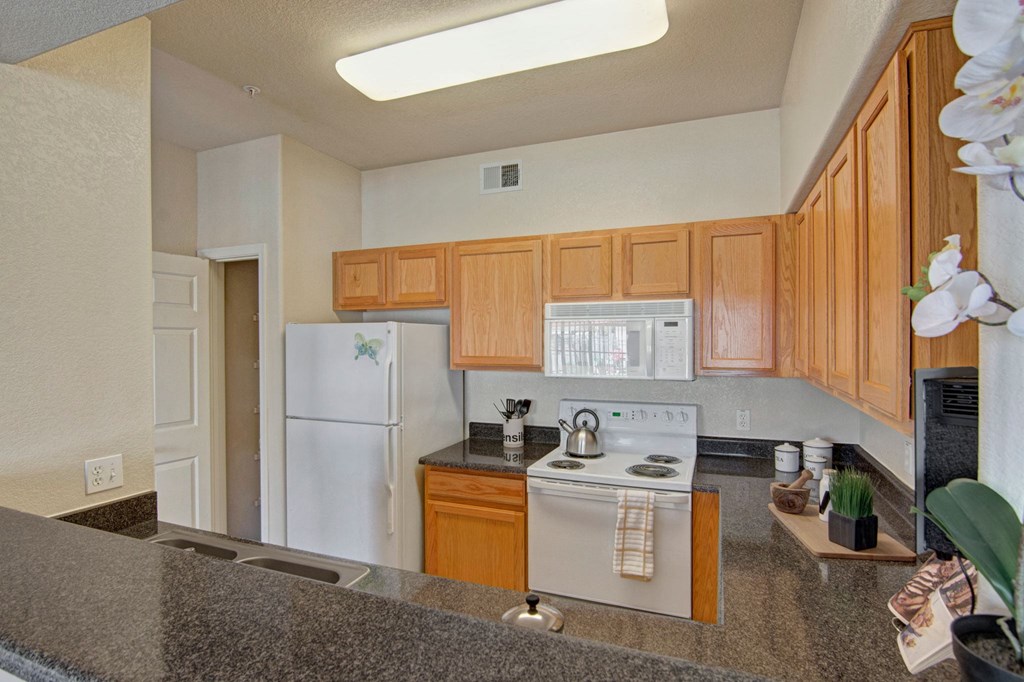 Cozy kitchen here at Dobson Towne Centre Apartments with light wood cabinets, white appliances, and a granite countertop. A kettle sits on the stove; orchids and decor add warmth.