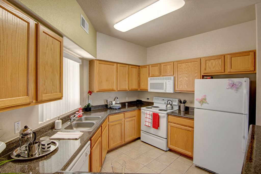 Modern kitchen here at Dobson Towne Centre Apartments with light wood cabinets, gray granite countertops, and white appliances. A red towel hangs on the oven, and two butterfly magnets adorn the fridge, creating a warm and inviting atmosphere.