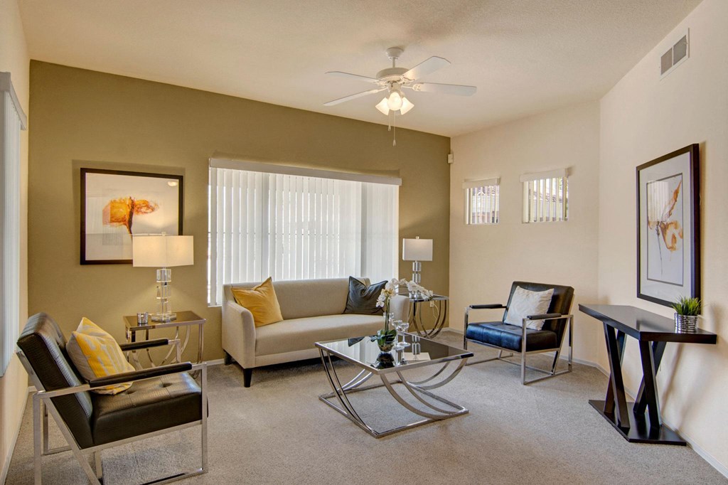 Modern living room here at Dobson Towne Centre Apartments with a neutral tone, featuring a beige sofa, two black chairs, glass coffee table, abstract art, and natural light from large windows.