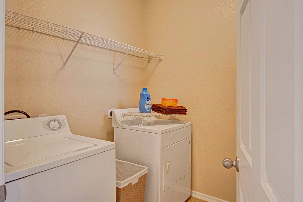 Compact laundry room here at Dobson Towne Centre Apartments with a white washer and dryer. Detergent and towels rest on the dryer. Beige walls and wooden floor create a clean, functional space.