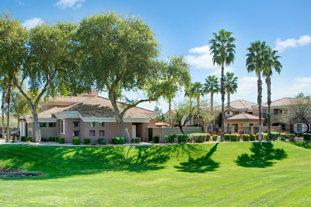 A serene suburban scene here at Dobson Towne Centre Apartments with a tan stucco house surrounded by lush green lawns, palm trees, and clear blue skies. Shadows create a tranquil atmosphere.