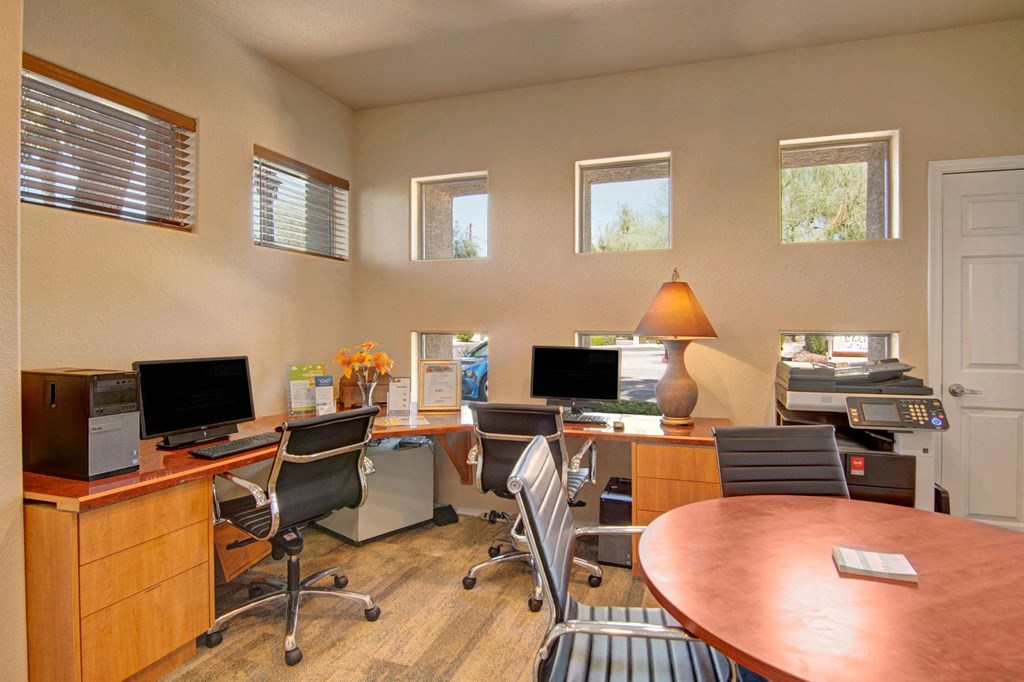 Modern office space here at Dobson Towne Centre Apartments with neutral tones, featuring two desks with computers, black chairs, a lamp, and a printer. Natural light streams through small windows.