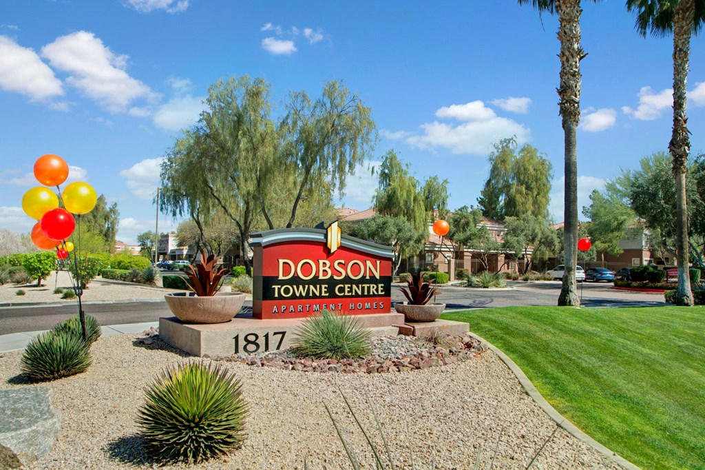 Sign for Dobson Towne Centre Apartment Homes surrounded by desert landscaping, balloons, and palm trees under a clear blue sky.