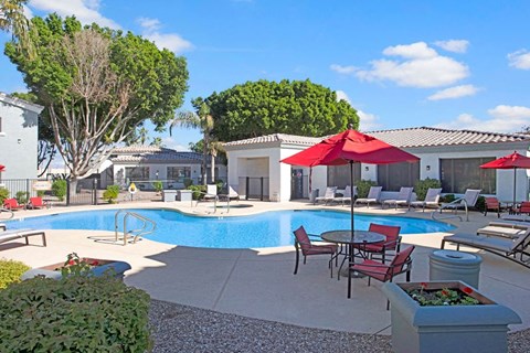 Sunny pool area here at Pala Mesa Apartments with turquoise water, surrounded by white lounge chairs. Red umbrellas provide shade. Trees and white buildings in the background. Relaxing vibe.