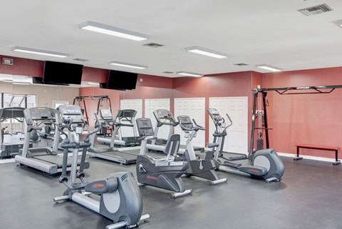 A modern gym here at Pala Mesa Apartments with various cardio machines, including treadmills and ellipticals, arranged on a dark floor. The room has white walls and two wall-mounted TVs.