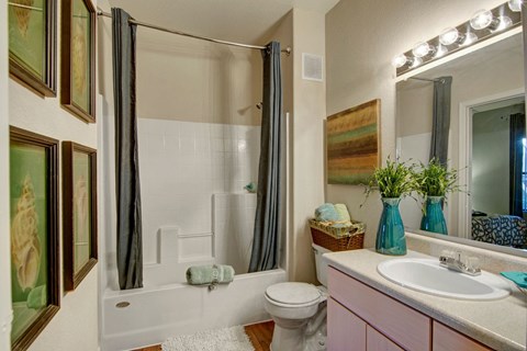 Cozy bathroom here at Pala Mesa Apartments with white fixtures, gray shower curtain, and pine cabinets. Wall art and potted plant add color. Soft lighting creates a warm ambiance.