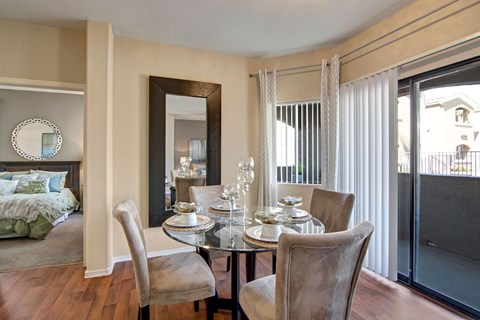 Dining area here at Pala Mesa Apartments with a glass table set for four, featuring beige velvet chairs. Large mirror and sliding doors brighten the space, leading to a cozy bedroom.