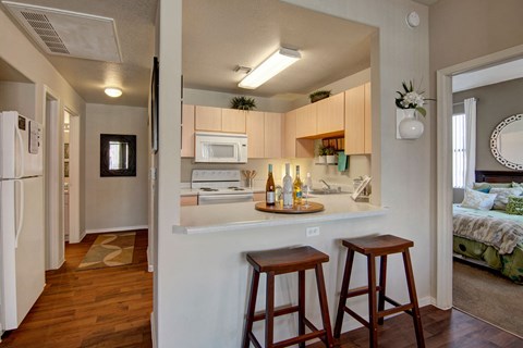 Compact kitchen here at Pala Mesa Apartments with light wood cabinets, a white stove, and microwave. Counter with wine bottles and glasses, two wooden stools. Cozy, inviting feel.