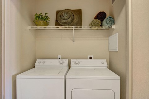 Compact laundry room here at Pala Mesa Apartments with a washer and dryer side by side. Above, a shelf holds a potted plant, three rolled towels, and spiral decor. Neutral tones.