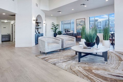 A modern, spacious resident lounge here at Pala Mesa Apartments with light wood flooring and two beige armchairs around a round marble table. Large plants, abstract art, and soft lighting create a tranquil, inviting atmosphere.