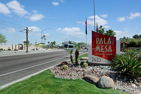Landscape view of Pala Mesa Apartment Homes sign with palm trees and desert plants. A streetcar passes by on a sunny day, blue sky with clouds.