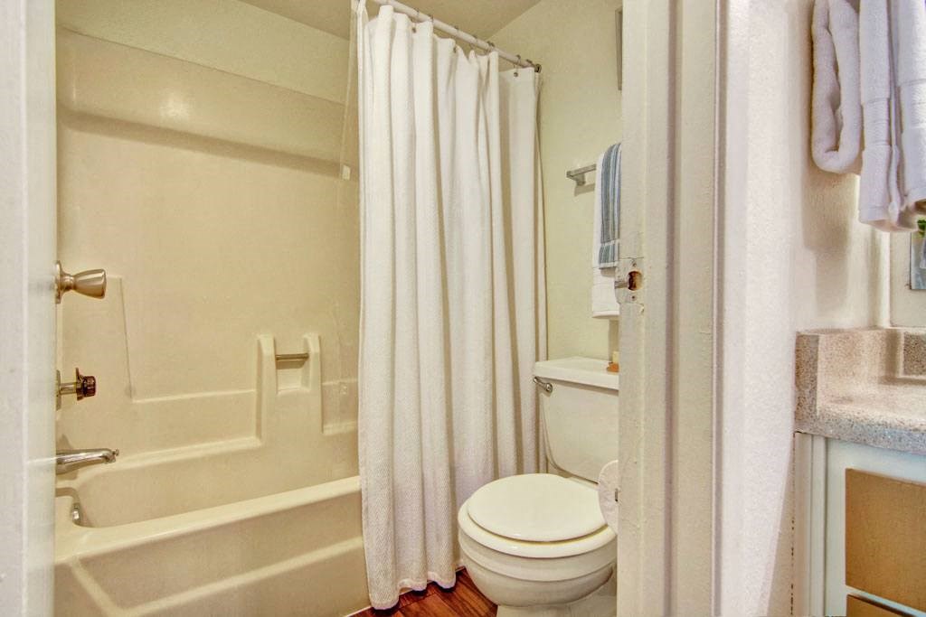 A small bathroom here at Paradise Falls Apartments with a white shower curtain over a bathtub, a toilet beside it, neatly folded towels on a rack, and a light countertop, conveying a clean and simple ambiance.