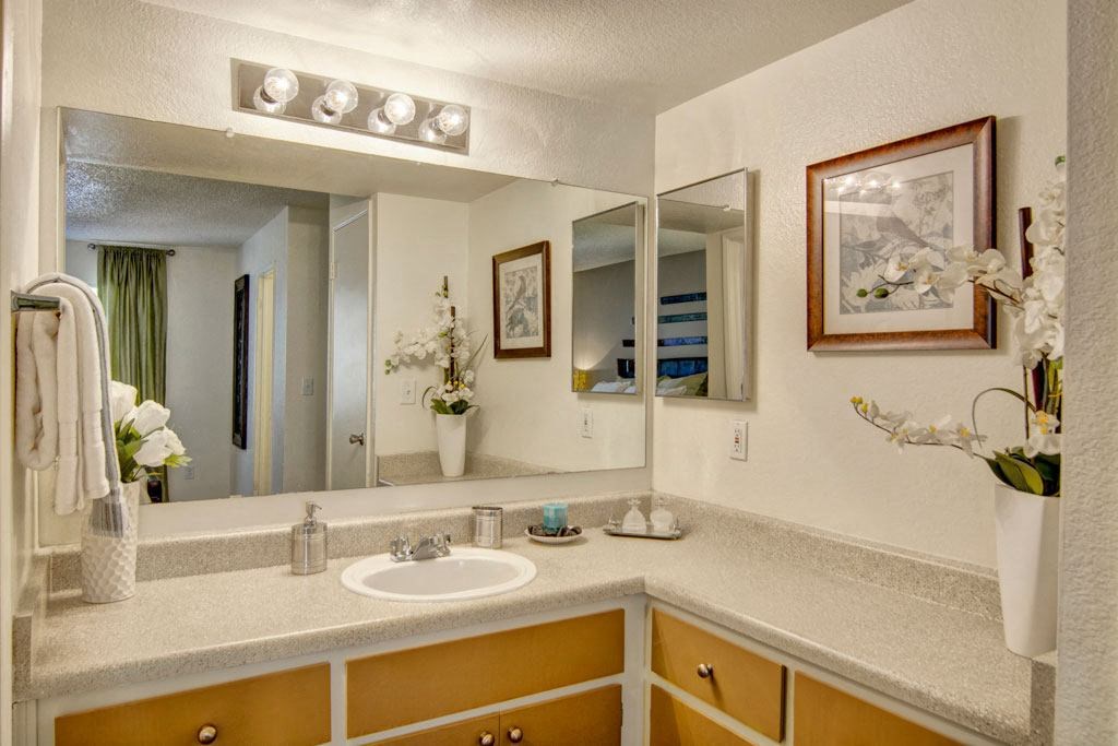 Bright bathroom here at Paradise Falls Apartments with a large mirror above a beige countertop. Decor includes white orchid flowers in vases and framed wall art, creating a serene ambiance.