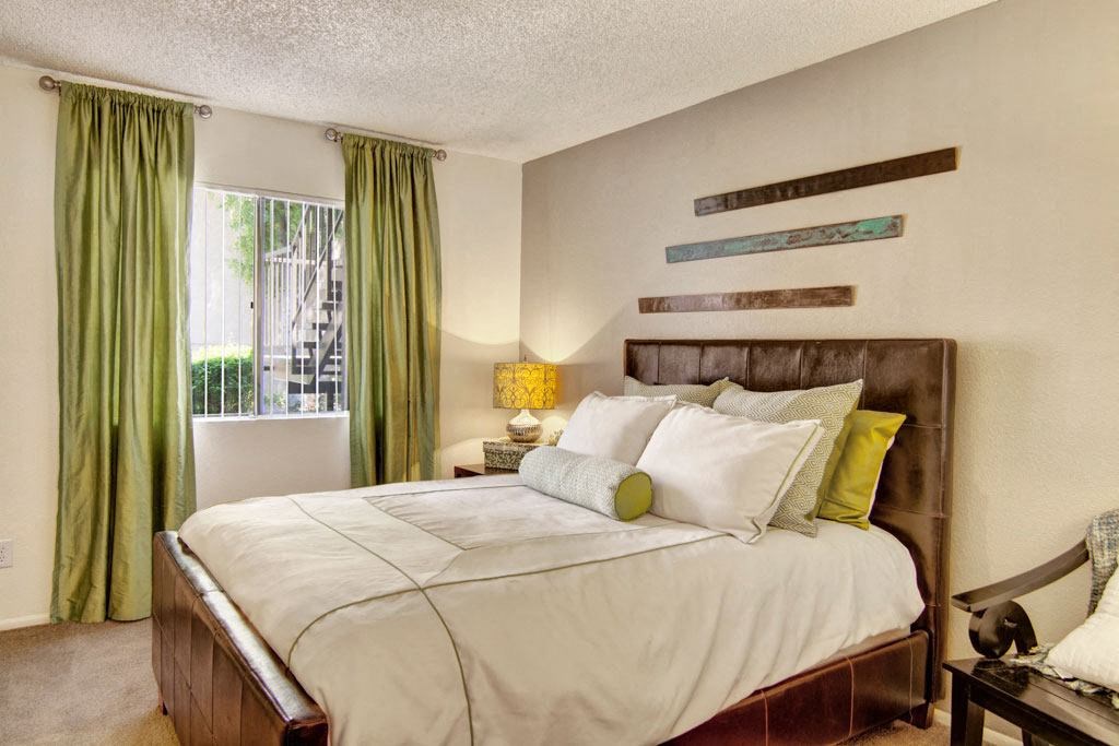 Cozy bedroom here at Paradise Falls Apartments with a brown leather bed, green and white pillows, and a matching bedspread. Green curtains cover a window. Wall decor adds elegance.