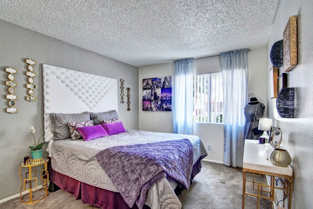 Cozy bedroom here at Paradise Falls Apartments with a modern gray and purple color scheme. Features a tufted white headboard, decorative mirrors, art, and a dress form. Soft, natural light filters through sheer curtains, creating a serene ambiance.