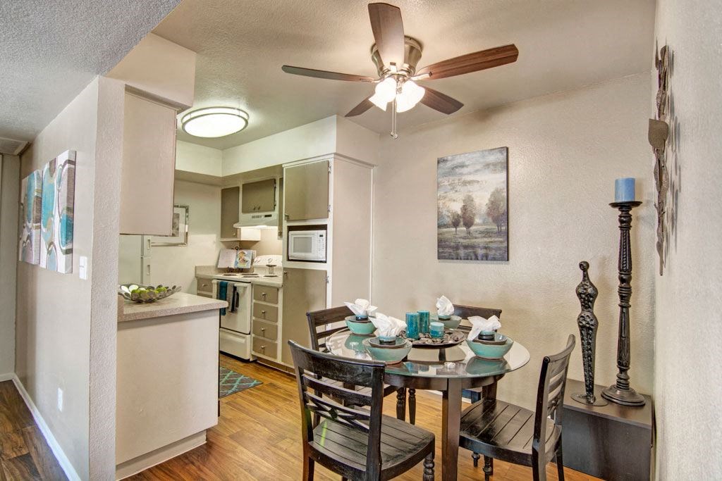 Cozy dining area here at Paradise Falls Apartments with a round table set for four, featuring teal dishes and napkins. Nearby, a modern kitchen and wall art create a warm, inviting atmosphere.
