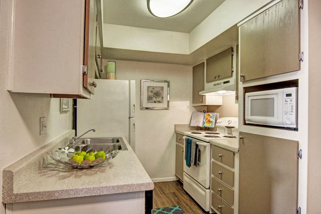 A small kitchen here at Paradise Falls Apartments with beige cabinets, a white stove, and a microwave. A bowl of green apples sits on the counter. The space feels cozy and functional.