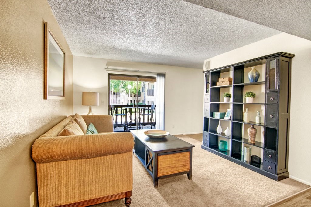 Cozy living room here at Paradise Falls Apartments with a beige sofa, wooden coffee table, and large black bookshelf. Sliding glass door opens to a patio, creating a bright and airy ambiance.