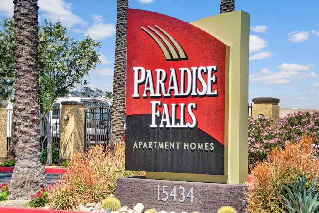 Red and beige sign for Paradise Falls Apartment Homes, surrounded by palm trees and shrubs, with a bright blue sky in the background.