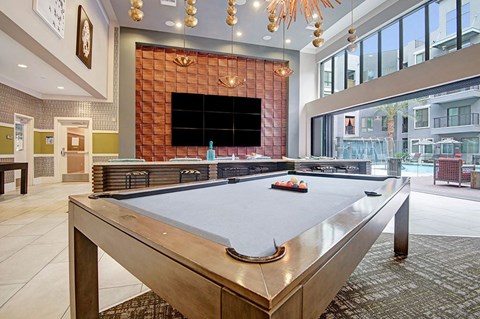 Modern entertainment room here at Park on Central Apartments with a pool table and colorful balls in the foreground. Large TV wall, contemporary art, and bright windows revealing a pool area.