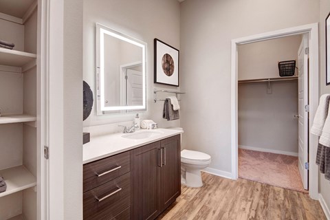 Modern bathroom here at Park on Central Apartments with wood flooring, a dark wood vanity, and a bright mirror above the sink. An open closet with a basket is visible in the background.