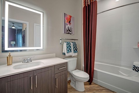 Modern bathroom here at Park on Central Apartments with glowing mirror over a sleek sink and wood cabinet. Maroon shower curtain next to a toilet with colorful towels above. Calm ambiance.