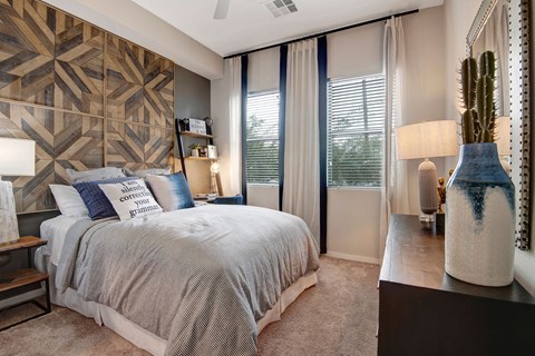 Modern bedroom here at Park on Central Apartments with geometric wood accent wall, cozy bed with gray and blue pillows, large window with sheer curtains, and a stylish lamp on a dresser.