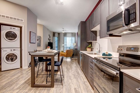 Modern open-concept kitchen and living area here at Park on Central Apartments. Grey cabinets, stainless steel appliances, stacked washer-dryer, and a wood dining table create a cozy feel.