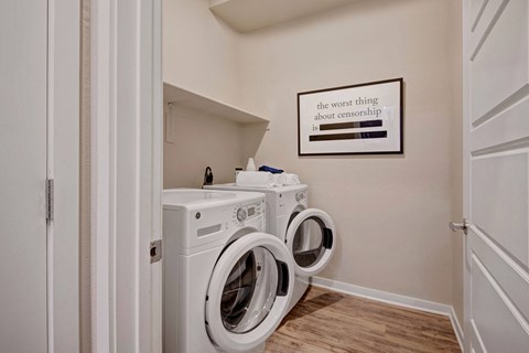 A small laundry room here at Park on Central Apartments with a washer and dryer, both doors open. Wall art with the phrase "the worst thing about censorship is" partially redacted. Clean and minimalistic.