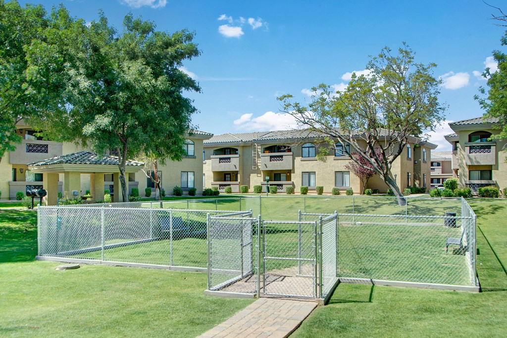 Spacious dog park here at Reserve at Arrowhead Apartments with chain-link fencing in a well-kept apartment complex. Surrounded by trees, green lawns, and beige buildings under a blue sky.
