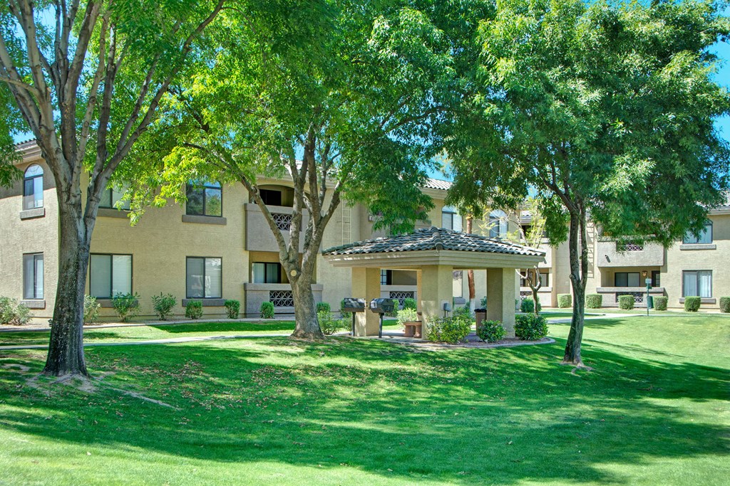 Sunny courtyard with green lawns, tall trees, and beige Reserve at Arrowhead Apartment complex featuring balconies. A shaded gazebo with a grill and seating is central.