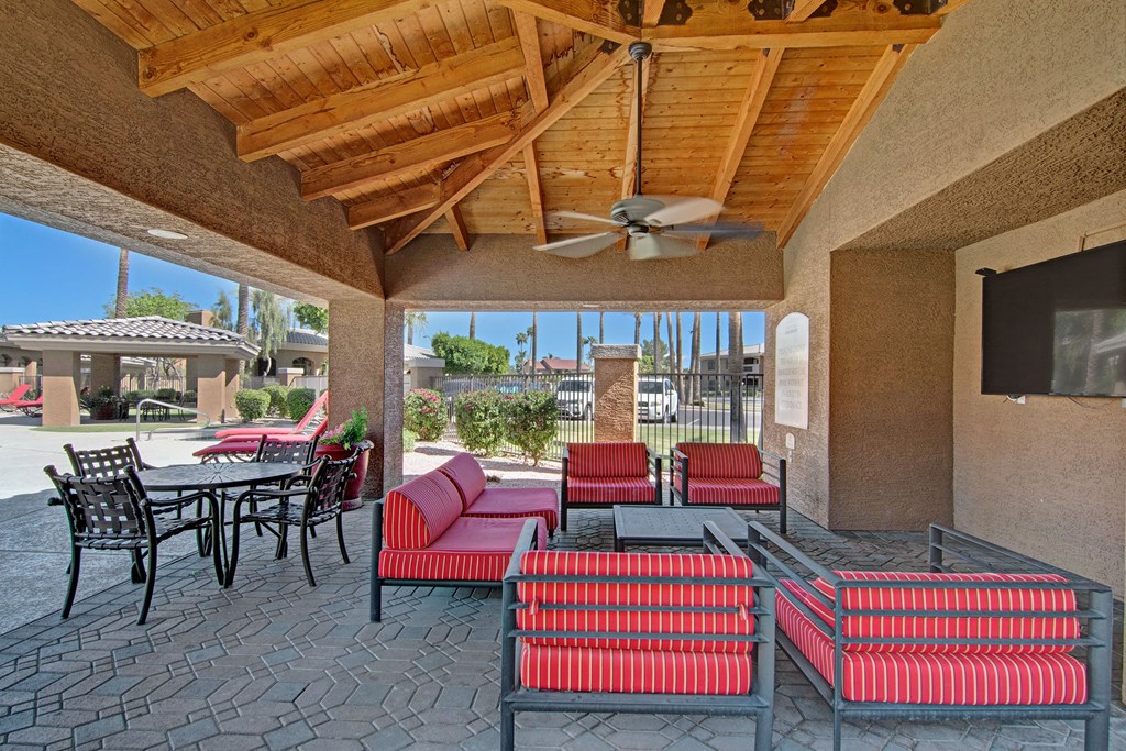 Outdoor patio here at Reserve at Arrowhead Apartments with red-striped seating beneath a wooden pergola. Black metal dining set, ceiling fan, TV, and view of sunny garden with palm trees. Relaxing ambiance.