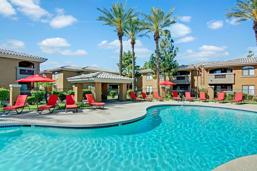 Reserve at Arrowhead Apartment complex pool area with clear blue water, surrounded by red lounge chairs and umbrellas. Palm trees and beige buildings create a relaxing atmosphere.
