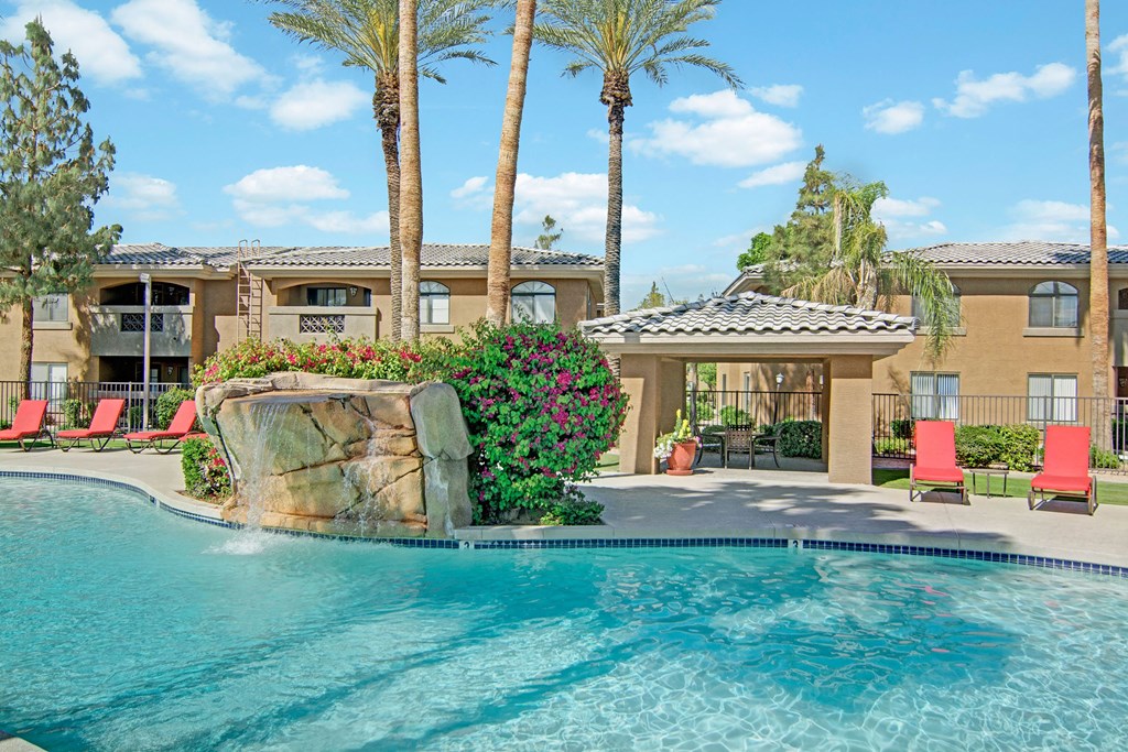 Outdoor pool area here at Reserve at Arrowhead Apartments with a rock waterfall feature, surrounded by palm trees and vibrant flowers. Red lounge chairs line the poolside, set against sunny blue skies.