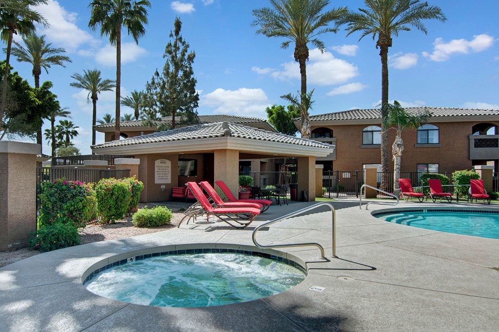 Outdoor relaxation area here at Reserve at Arrowhead Apartments with a hot tub and pool, surrounded by palm trees and red lounge chairs under a clear blue sky, conveying a tranquil vibe.