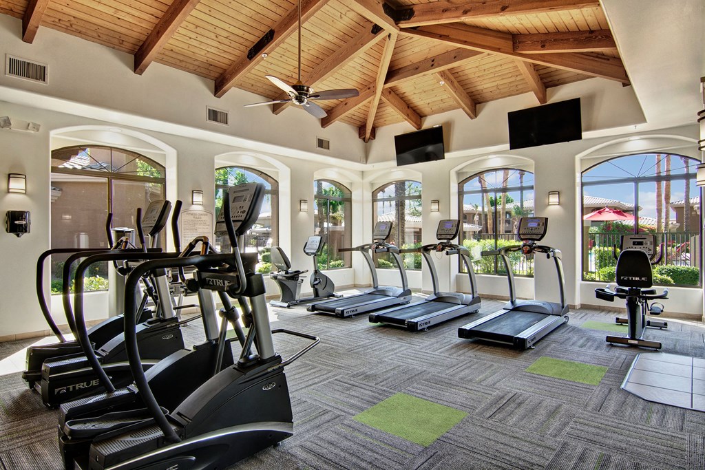 Spacious gym here at Reserve at Arrowhead Apartments with a vaulted wooden ceiling, featuring ellipticals, treadmills, and a stationary bike. Large windows offer a view of greenery outside.