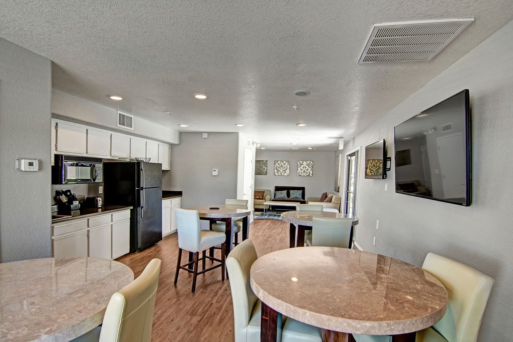 Bright Kitchen and dining room here at Reserve at Arrowhead Apartments with cream walls and wood floors, featuring round tables with beige chairs. A kitchen is visible, and a wall-mounted TV is on the right. Calm ambiance.