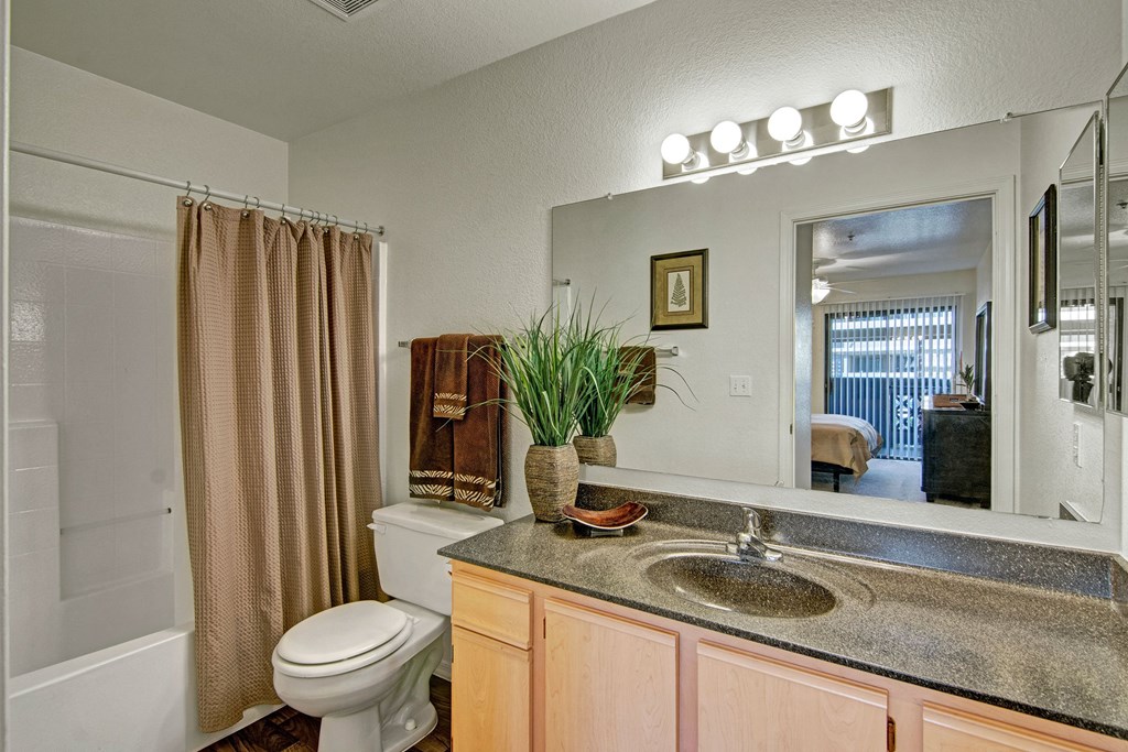 A bathroom here at Reserve at Arrowhead Apartments with a dark countertop, light wood cabinets, and a white sink. Brown towels hang near a plant. A door leads to a bedroom with a large window.