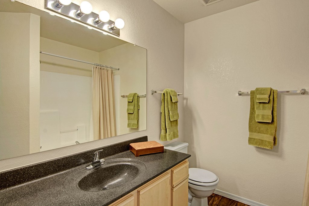 A bathroom here at Reserve at Arrowhead Apartments with a large mirror, bright overhead lights, and a dark countertop with a sink. Green towels hang neatly, with a beige shower curtain in the background.