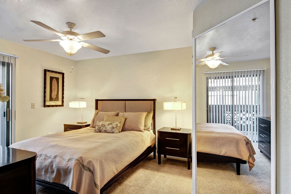 Cozy bedroom here at Reserve at Arrowhead Apartments with a beige bedspread, dark wood furniture, and soft lighting. Ceiling fans and mirrored closet reflect a calming, relaxed ambiance.
