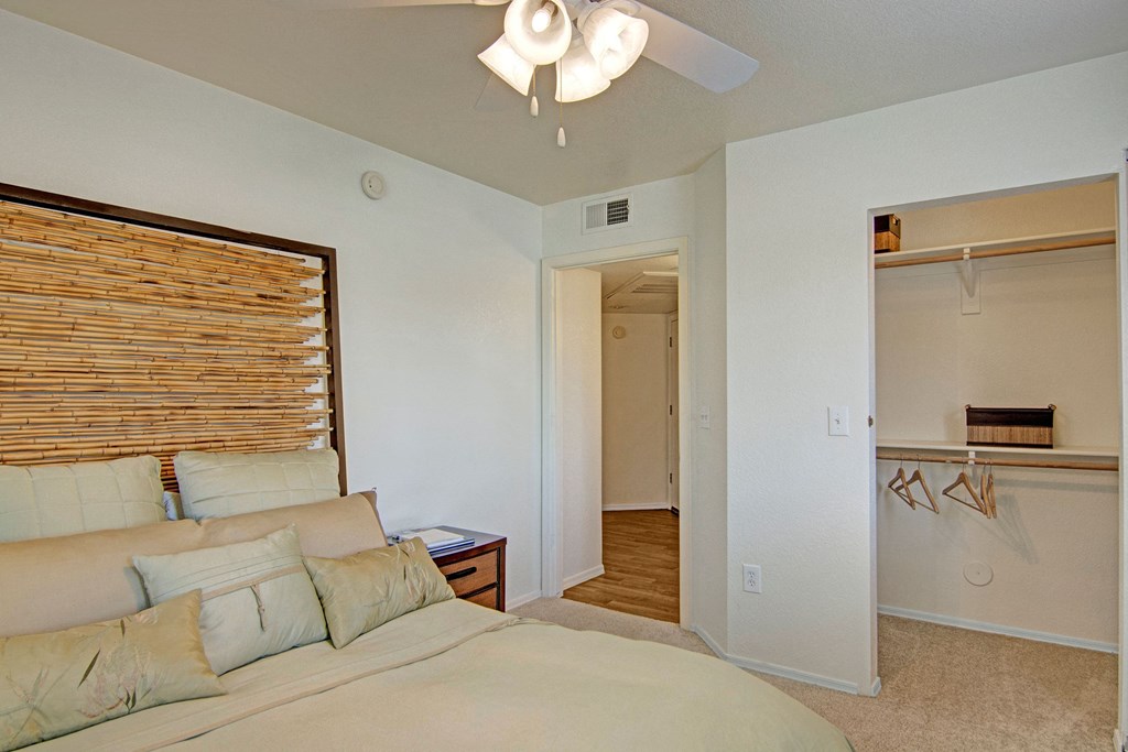 Bedroom here at Reserve at Arrowhead Apartments with a beige bed featuring multiple cushions and a bamboo headboard. An open closet with wooden hangers is on the right, conveying a cozy atmosphere.