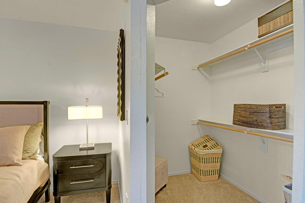 A cozy bedroom corner here at Reserve at Arrowhead Apartments with a black nightstand beside a bed with light bedding. A lit lamp is on the nightstand. Adjacent, an organized walk-in closet with shelves, baskets, and hangers.