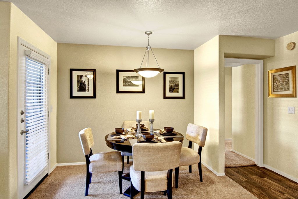 Cozy dining area here at Reserve at Arrowhead Apartments with beige walls, round table set for four with brown dishes. Framed photos on walls, soft lighting, and a door with blinds.