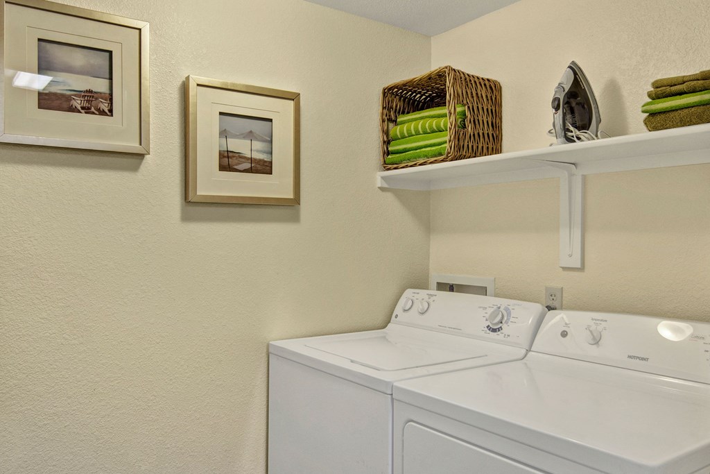 Laundry room here at Reserve at Arrowhead Apartments with white washer and dryer, beige walls, beach-themed framed photos. Shelf holds green towels, iron, and a wicker basket. Cozy ambiance.