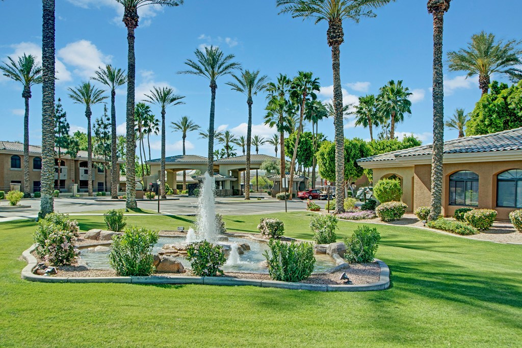 Lush landscape here at Reserve at Arrowhead Apartments with tall palm trees and a central fountain on green lawns. Buildings with tiled roofs and arched windows form a serene, resort-like setting.
