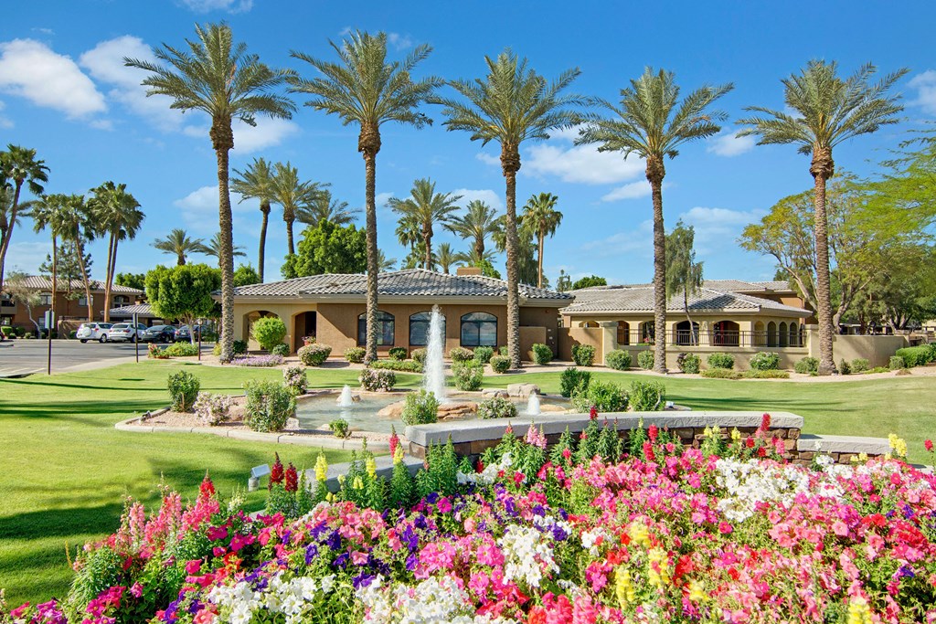 Lush garden with vibrant flowers in the foreground, a small fountain, and palm trees lining two Mediterranean-style buildings here at Reserve at Arrowhead Apartments under a clear blue sky.
