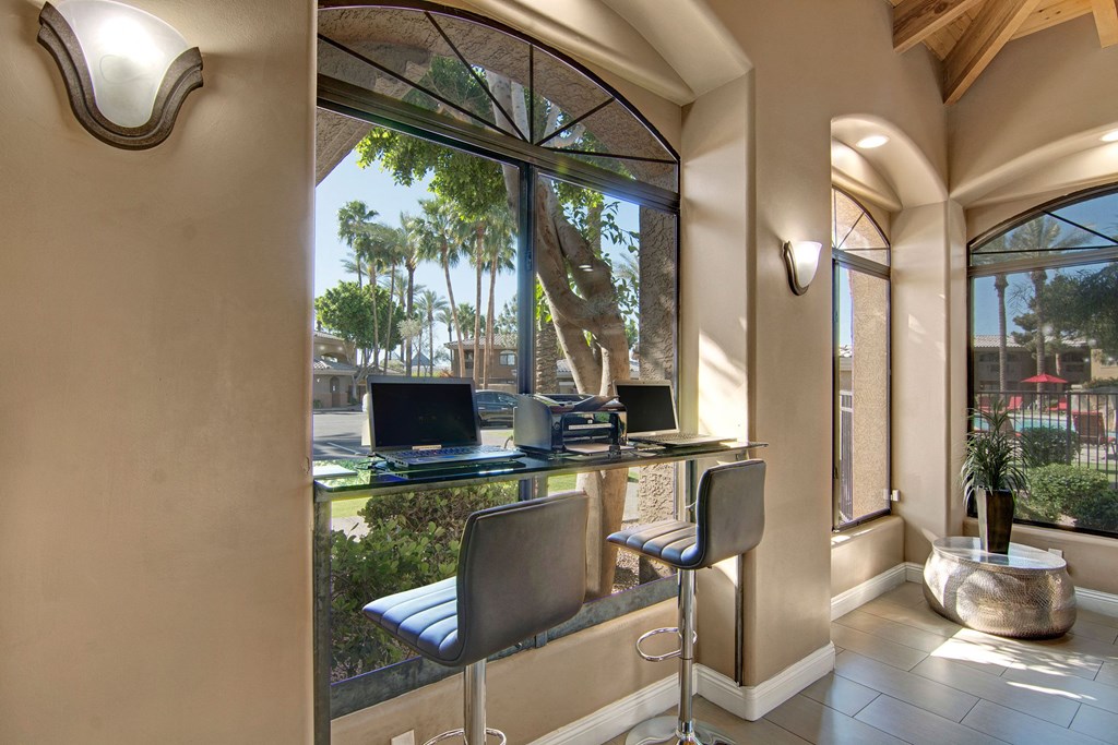 Bright, modern workspace here at Reserve at Arrowhead Apartments with two laptops on a glass bar table, high stools, large windows showing palm trees outside, and soft beige walls.